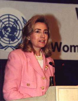    Hillary Rodham Clinton wearing pink suit giving speech at a Woman's Day conference wearing the Illuminati Phoenix Pin.   